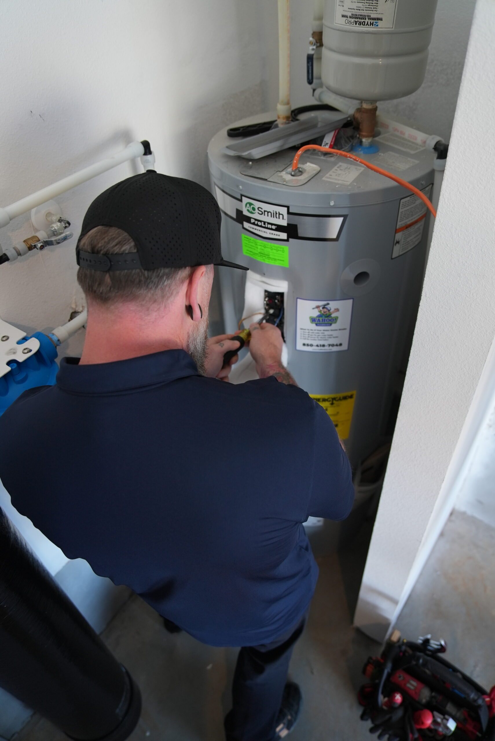 Wahoo Plumbing technician working on a residential water heater system, seen from an over-the-shoulder angle wearing a company polo and black cap.